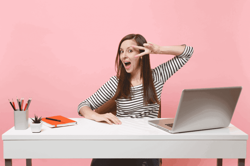 young-excited-specialist-woman-with-opened-mouth-showing-victory-sign-white-desk-with-contemporary-laptop